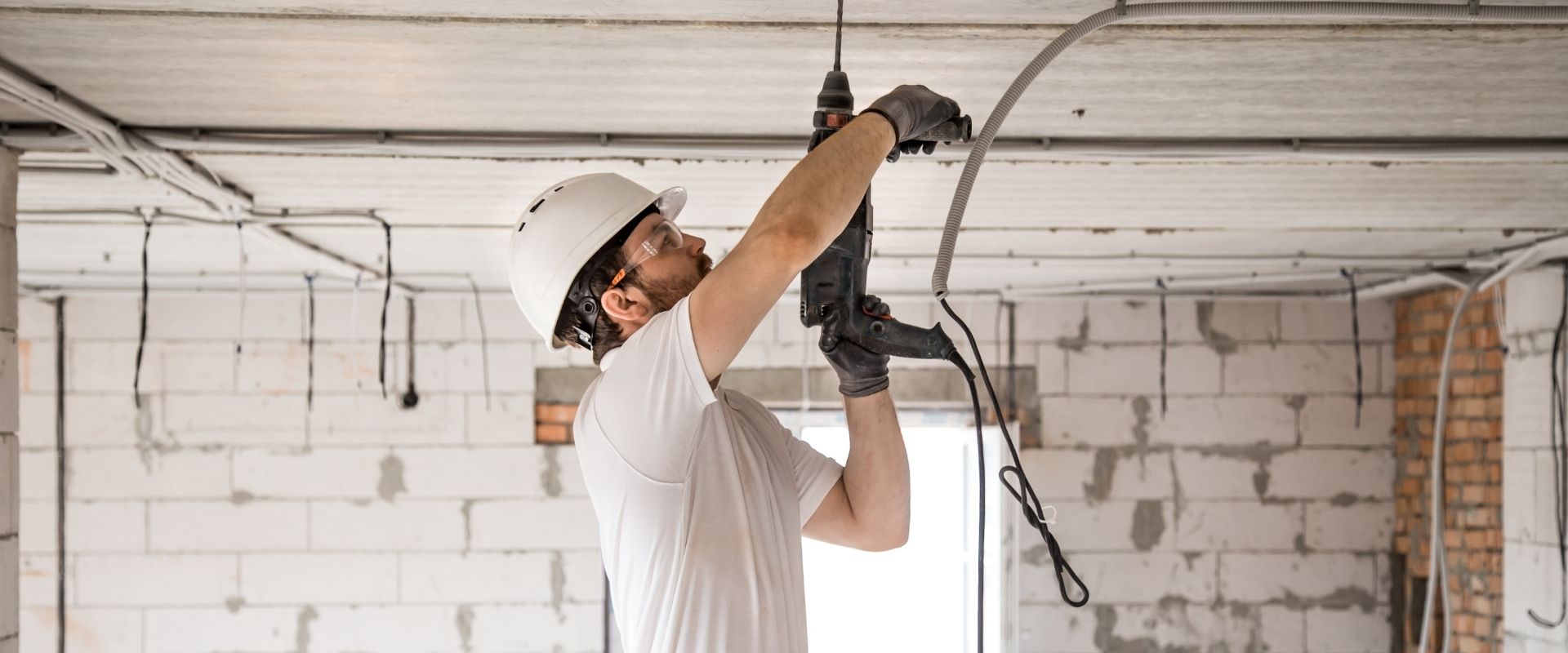 A person wearing a hard hat and gloves uses a drill on a ceiling in a room under construction with exposed wiring and cinder block walls.