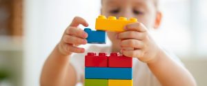 A child stacking colorful toy blocks, adding a yellow block to a blue one on top of a red, blue, green, and orange tower.